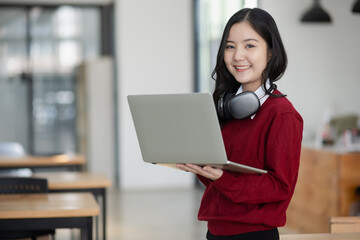 Happy young asian woman wearing earphones and using laptop computer at desk in office.