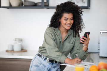 latin young woman stands holding smartphone taking notes in kitchen