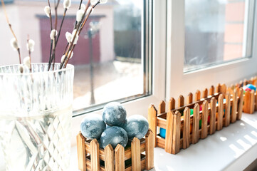 Vase with willow and colored Easter eggs in a wooden box on the windowsill near the window