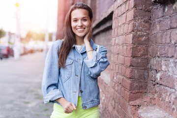 Happy young woman wearing denim jacket laughing looking at camera standing on street. Smiling teen generation z hipster girl posing outdoor.