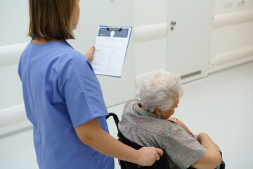 Nurse pushing senior patient in a wheelchair, reading medical document in clipboard. Emotional...