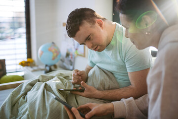 Young man with down syndrome sitting in bed with mom, looking at smartphone in morning. Morning...