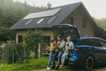 Family with electric car standing in front their house with solar panels on roof. Solar energy and...