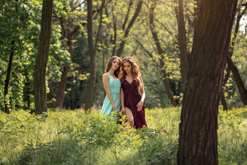 Photo of fashionable twins models with perfect make-up and hairstyle standing in forest on green grass meadow with yellow flowers. Spring beauty concept. Shot of two gorgeous women in evening dresses