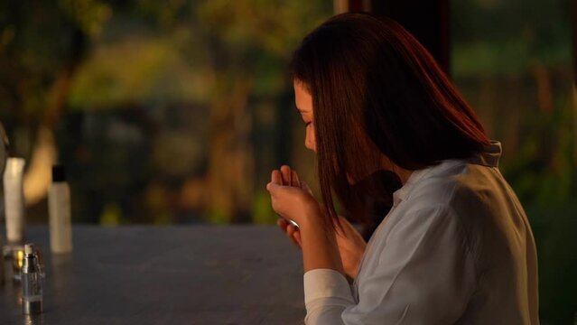 Asian young woman takes care of her skin in the kitchen using skin gel against the background of a large window during sunset. Asian young woman sniffing skin gel.