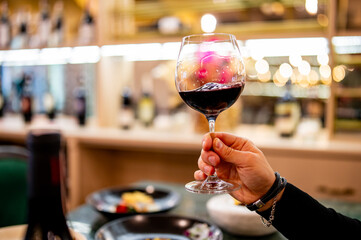 A hand holds a half-full glass of red wine against a blurred restaurant backdrop, exuding an elegant dining atmosphere. The warm lighting and reflections on glass surfaces suggest an upscale setting