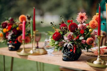 Variegated multi-colored flower arrangements in vases in the shape of a skull, Mexican style in the decor. On the festive table in the wedding banquet area, compositions of flowers and greenery, candl