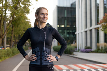 A fit and happy woman takes a break from running, her hands on her hips as she pauses to enjoy the city scenery. Earphones in, she's dressed in athletic gear, ready to continue her fitness routine.