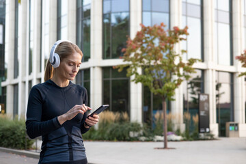 A focused woman enjoying her playlist while handling her smartphone in a leisurely stance, dressed in fitness attire, with modern architecture and greenery in the background.