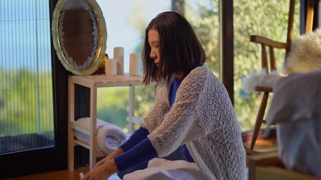 Asian young woman folding towels at home and looking at herself in the mirror while sitting on the floor against the background of a large window