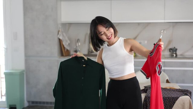 Happy young Asian girl trying to choose a dress standing by table in the kitchen