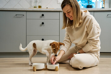Woman putting food bowl with feed for her dog on the floor in kitchen, Female owner spending time together with pet at home, Animal feeding and pet care