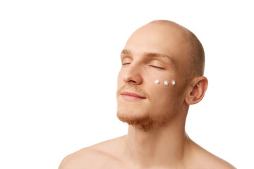 Fototapeta premium Portrait of handsome bald young man standing with eyes closed with moisturizing cream on face isolated over white studio background. Concept of skin care, male beauty and cosmetics, youth, self-care