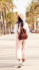 Rear view of a woman traveling walking alone. Nomadic people outdoors walking in a coast full of palm trees, during summertime. Solo trip. Empowered woman. Full length vertical image.