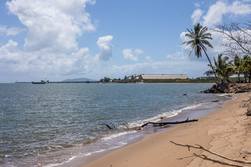 A tropical beach with palm trees, calm water and a sugar terminal with an extremely long sugar export jetty under a blue and cloudy sky at Lucinda in tropical Queensland, Australia.