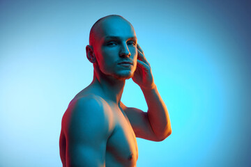 Portrait of handsome young bald man posing shirtless, looking at camera against blue studio background in neon light. Concept of male beauty, sportive and healthy lifestyle, body care