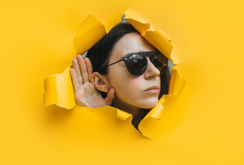 Close-up of a woman's head in black glasses and ear with her hands through a hole in the paper....