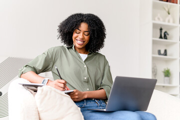 A content African-American woman engages in multitasking, writing in a notebook while...