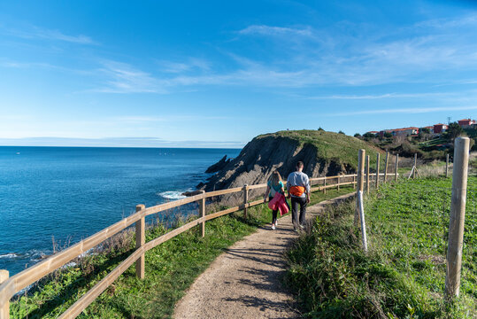Couple of hikers with their backs turned walking along a mountain path on the coast of the Basque Country next to the tourist town of Bermeo on a sunny day.