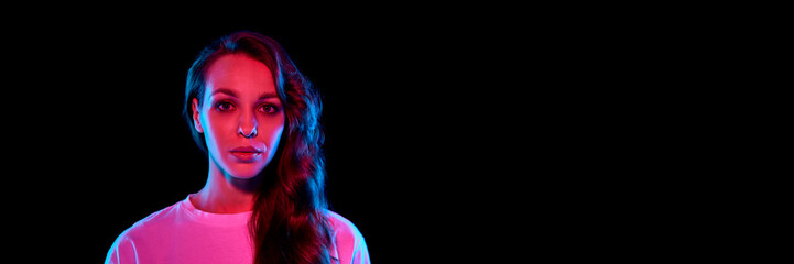 Portrait of beautiful young woman with brunette curly hair looking at camera against black background in neon. Concept of youth, lifestyle, fashion, emotions. Banner. Empty space to insert text, ad