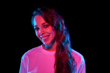 Happiness. Portrait of beautiful young woman with curly hair posing with smile against black studio background in neon light. Concept of youth, lifestyle, casual fashion, human emotions