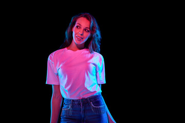 Portrait of beautiful young woman in white t-shirt and jeans posing against black studio background in neon light. Concept of youth, lifestyle, casual fashion, human emotions