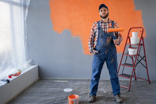 Handsome male worker in cap and overalls with paint roller in empty room on orange wall background.