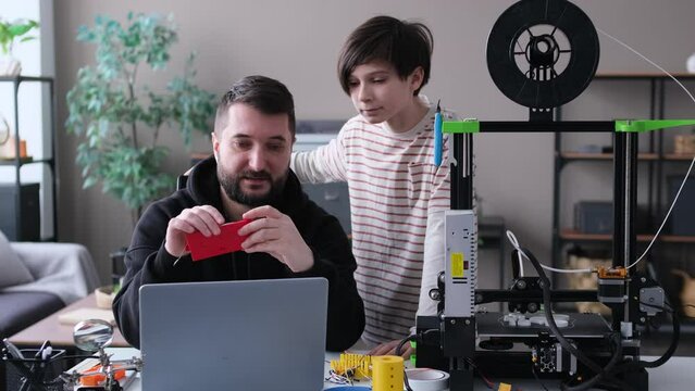 Positive Caucasian son and father at home work shop, engaged in constructing project researching using laptop and 3D printer. Family bonding and teamwork concept.