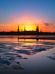 Naklejka premium A beautiful, colorful sunrise landscape with city centre building silhouette against bright morning sky. A winter lansdcape with frozen river Daugava in Latvia capital Riga.