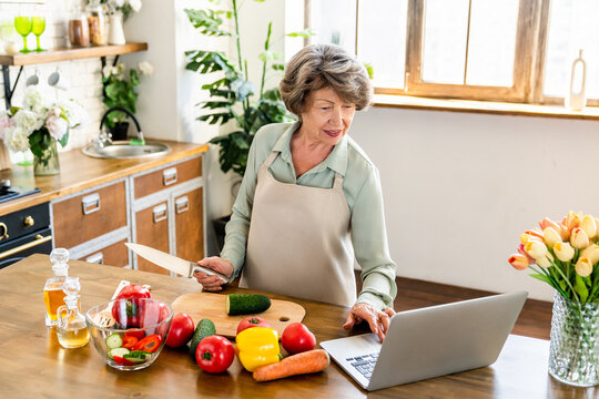 Modern senior grandmother cooking vegetable salad while using laptop to find recipe. Housewife old lady elderly woman in apron cutting vegetables for lunch and vlogging online