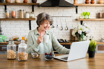 Breakfast at home. Grandmother drinking hot tea coffee beverage while browsing on laptop, doing online shopping. Healthy eating habits concept - obrazy, fototapety, plakaty