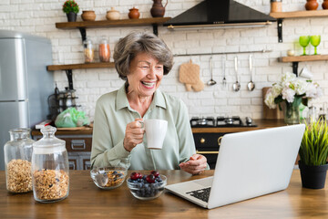Laughing happy old elderly woman watching movie video on laptop at home kitchen while having healthy breakfast. Grandmother drinking hot beverage - obrazy, fototapety, plakaty