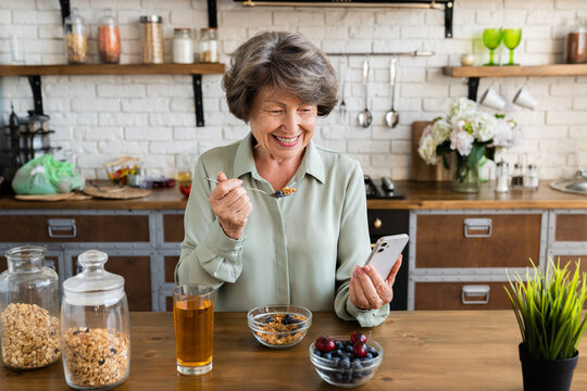 Smiling caucasian senior woman eating healthy breakfast while using smart phone for social media, online shopping at home kitchen. Grandmother eating oat cereals in the morning