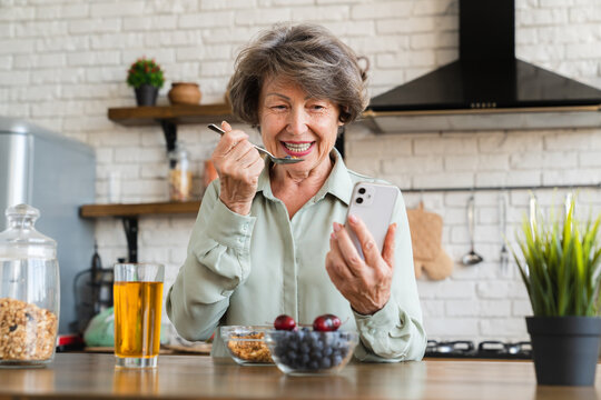 Cheerful caucasian senior woman eating oat cereals for breakfast while browsing smart phone, doing online shopping from home. Healthy diet eating habits