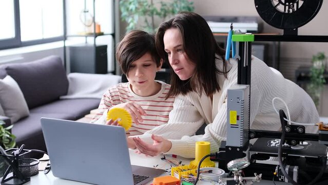Cheerful son and mother study a new technology together. Exploring 3D printer and his plastic products, engaged in education process. Family bonding and teamwork concept.