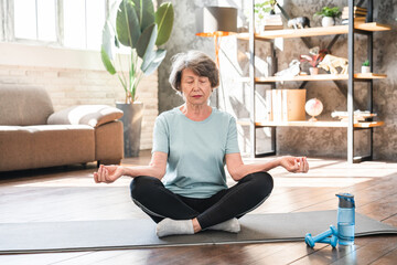 Serene people concept. Caucasian fit elderly athlete woman in sporty outfit meditating on fitness mat. Balance and calmness, yoga classes at home.