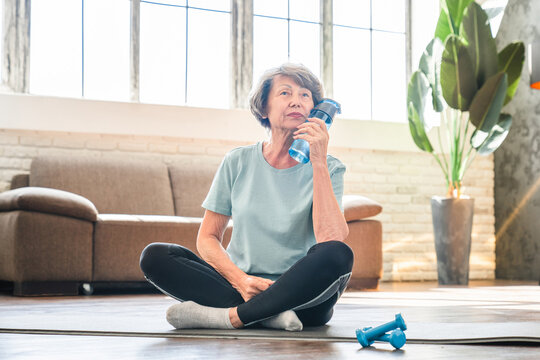 Tired exhausted caucasian senior woman relaxing after training on fitness mat in lotus position. Grandmother drinking water after home workout