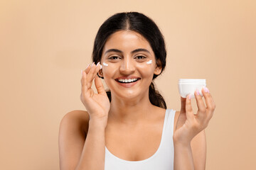 Indian Female Applying Moisturising Cream On Face While Standing Against Beige Background