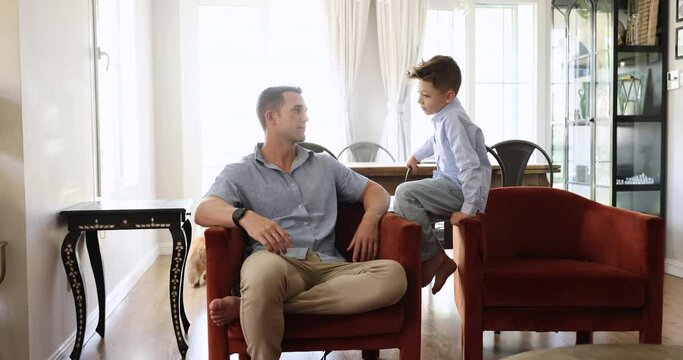 Young loving father and little cute son lead carefree tattle seated on cozy armchair in living room, spend time together, enjoy talk, share news, asking and giving advice. Child upbringing, fatherhood