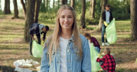 Portrait of volunteer young woman looking at camera near volunteers from eco team which with plastic bags collecting trash in the park. Society against pollution.