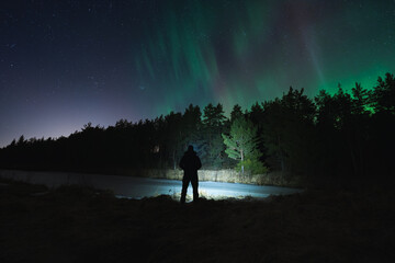 Night scene, nature of Estonia. Silhouette of a man with a flashlight in a dark forest with a starry sky and northern lights.
