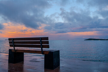 A wooden bench positioned on a dock offers a serene spot to admire the natural landscape as the sun sets over the ocean, painting the sky with an array of colors