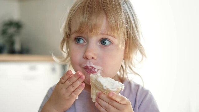 Baby girl enjoying ice cream. Pretty little toddler eating an ice-cream indoors, at home. Dining room background. Small child eats plombir and cream messy on her mouth. Cute kid with tasty sweet food.