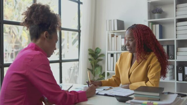 Two African American business women are talking in a private office, they are discussing about project that their boss ordered them to finish before the presentation day.