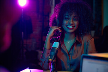 Naklejka na ściany i meble Smiling young African woman, social media influencer, blogger sitting with microphone at podcast interview. Making show. Concept of online communication, modern technology, mass media