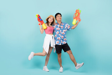 Full body shot of happy young asian couple in summer outfits with water gun and looking at camera in blue background.