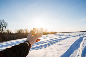 Hand holding cup of coffee on the background of the glare of the sun, winter landscape, drinking tea in nature, winter evening in the forest.