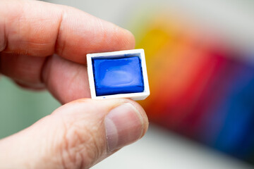 A hand showing a mini blue watercolor cube with a multicolored watercolor background. Landscape format.