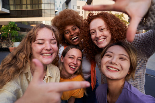 Happy selfie of an internacional group of female friends making frame gesture with fingers outdoors in the city. Young women looking at camera smiling.