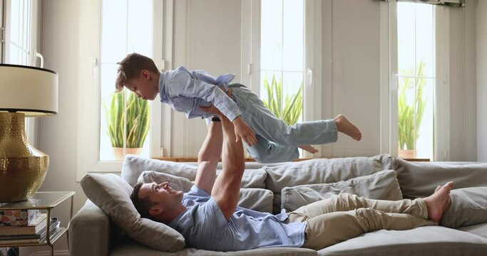 Cheery father plays with little son, lifting lovely kid on outstretched hands, enjoy playtime in cozy living room. Preschooler boy have fun with dad, imagining flying in the air, pretend to be a plane
